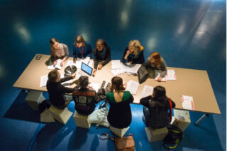 Eine Gruppe von Menschen sitzt um einen rechteckigen Tisch herum und ist in eine Diskussion vertieft. Vor ihnen liegen Papiere, Notizbücher und ein Laptop (von oben betrachtet).
