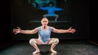 Ein Mann in einem weißen Outfit hockt mit ausgestreckten Armen auf einer Bühne, hinter ihm wird das Bild eines Kindes in einer ähnlichen Pose projiziert.