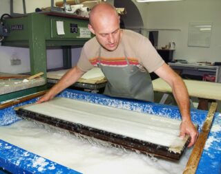 In einer Werkstatt verwendet ein Mann einen großen Holzrahmen, um handgeschöpftes Papier herzustellen, indem er es in einen Bottich mit Wasser und Zellstoff taucht.