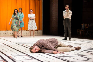 Im Vordergrund liegt ein Mann auf dem Boden, während im Hintergrund drei Frauen und ein Mann auf einer Bühne mit gelbem Vorhang stehen.