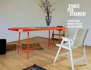 Ein minimalistischer Raum mit einem roten Tisch, einem weißen Stuhl, einer Vase mit Blumen, Holzboden und einem Text mit Namen und Veranstaltungsdetails an der Wand.