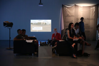 Eine Gruppe von Menschen sitzt auf Sofas in einem dunklen Raum und blickt auf eine Projektion an der Wand. Einige schreiben und eine Person benutzt ein Telefon.