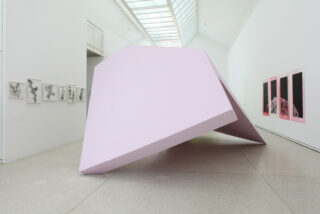 Große, blassrosa geometrische Skulptur, die schräg auf dem Boden einer Kunstgalerie mit weißen Wänden steht; im Hintergrund hängen gerahmte Kunstwerke an den Wänden.