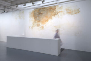 Eine verschwommene Figur bewegt sich hinter einer weißen Trennwand in einem minimalistischen Galerieraum mit einem strukturierten, abstrakten Wandkunstwerk in Braun und Beige.