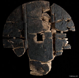 Zerbrochenes, altes Holzrad mit einem quadratischen Loch in der Mitte, aus mehreren dunklen Fragmenten zusammengesetzt, vor einem schwarzen Hintergrund dargestellt.