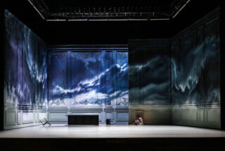 Eine Person sitzt allein in der Ecke eines großen, leeren, reich verzierten Raums, an dessen Wände dramatische Bilder eines Gewitterhimmels projiziert werden.