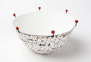 Eine weiße Keramikschale mit einem pixeligen schwarz-roten Muster und kleinen roten Würfeln, die am Rand mit dünnen schwarzen Stäben befestigt sind.