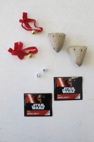 Zwei rote Bänder mit Glöckchen, zwei Ton-Okarinas mit Löchern, ein kleiner sechsseitiger Würfel und zwei ungeöffnete Star Wars Cosmic Shells-Packungen auf einer weißen Oberfläche.