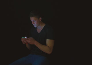 Ein Mann sitzt in einem dunklen Raum und schaut auf sein Smartphone, das sein Gesicht und seine Hände beleuchtet.