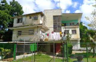 Ein zweistöckiges, verwittertes Wohnhaus mit Wäsche, die auf einem Balkon hängt, umgeben von einem Maschendrahtzaun und viel Grün.