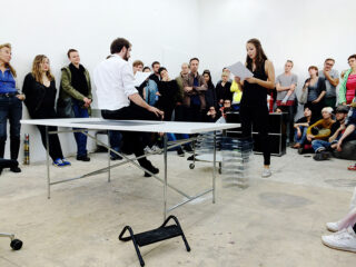 In einem Raum mit weißen Wänden und minimaler Möblierung steht eine Frau und liest einem sitzenden Mann vor einer Menschenmenge aus einer Zeitung vor.