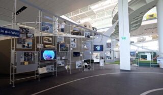 Ein Ausstellungsraum umfasst eine Metallausstellungsstruktur mit Fotos, Bildschirmen und Objekten zum Thema Technologie und Transport in einer modernen, gut beleuchteten Halle.