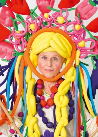 Eine ältere Frau in einem farbenfrohen Kostüm aus gedrehten Luftballons, inklusive Kopfschmuck und Strängen in verschiedenen leuchtenden Farben, posiert vor einem schlichten Hintergrund.