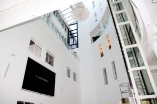 Das Innere eines modernen, mehrstöckigen Gebäudes mit weißen Wänden, Fenstern, einem Aufzugsschacht, einem großen schwarzen Bildschirm und einer kugelförmigen Deckenleuchte.