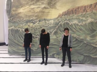 Drei Personen stehen mit gesenktem Kopf vor einer großen Kulisse, die stürmische Wellen und eine felsige Küste zeigt.