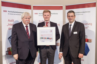 Drei Männer in Anzügen stehen im Innenbereich; der Mann in der Mitte hält eine gerahmte Urkunde in den Händen. Im Hintergrund sind Transparente mit dem Logo der Hochschulrektorenkonferenz zu sehen.