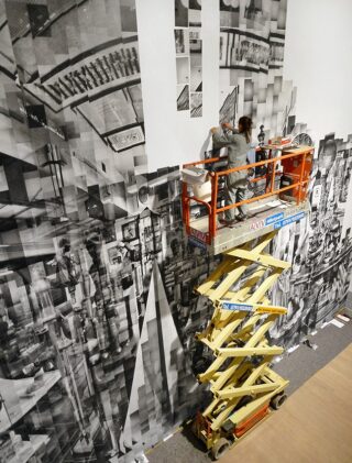 Zwei Personen installieren auf einer Scherenbühne ein großes Schwarz-Weiß-Collage-Kunstwerk an einer hohen Wand in einem Innenbereich.