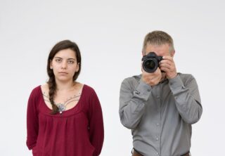 Eine Frau in einem roten Oberteil steht neben einem Mann in einem grauen Hemd, der eine Kamera vor sein Gesicht hält und vor einem schlichten weißen Hintergrund ein Foto macht.