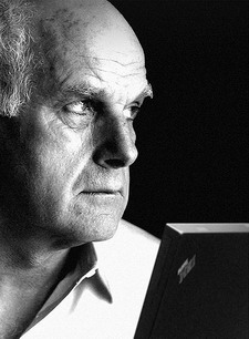 Schwarzweißfoto eines älteren Mannes mit ernstem Gesichtsausdruck, der nach links blickt und vor einem offenen Laptop sitzt.