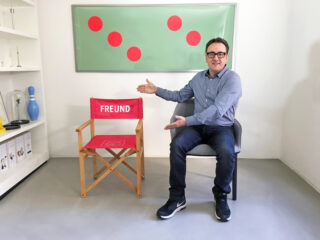 Ein Mann sitzt drinnen auf einem Stuhl und zeigt auf einen leeren Regiestuhl mit der Aufschrift „FREUND?“. An der Wand hinter ihm hängt ein gerahmtes grün-rotes Kunstwerk.