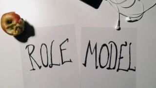 Zwei Blätter Papier mit den mit schwarzem Filzstift handgeschriebenen Worten „ROLLE“ und „MODELL“, auf einem Tisch platziert, zusammen mit einem angebissenen Apfel, Kopfhörern und einem Teil eines Smartphones.