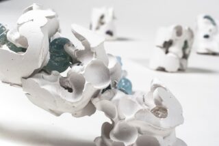 Nahaufnahme abstrakter weißer Keramikskulpturen mit eingebetteten hellblauen Glaselementen vor einem schlichten weißen Hintergrund, in der Ferne sind weitere Skulpturen unscharf zu sehen.