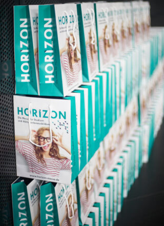 Auf schwarzen Regalen sind Reihen türkis-weißer Papiertüten mit der Marke HORIZON aufgereiht. Auf den Tüten ist das Foto einer lächelnden Person mit Brille abgebildet.