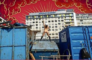 Eine Person mit Stiefeln und Helm steht mit erhobenem Arm auf einem Müllcontainer. Im Hintergrund sind ein großes Gebäude und rote Wolken abgebildet.