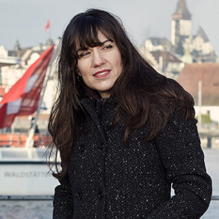 Eine Frau mit langem dunklem Haar und einem dunklen Mantel steht im Freien in der Nähe von Wasser, im Hintergrund sind Boote und Gebäude zu sehen.