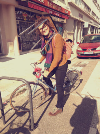 Eine Frau mit Brille, Schal und Rucksack steht neben einem geparkten Fahrrad auf einer Stadtstraße und lächelt in die Kamera.