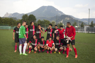 Eine Herrenfußballmannschaft in schwarz-roten Uniformen posiert für ein Gruppenfoto auf einer Wiese mit Bergen und Bäumen im Hintergrund.
