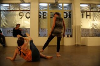 Zwei Personen führen in einem Raum mit großen Fenstern einen Tanz oder eine Aufführung durch, während zwei andere im Hintergrund sitzen und zuschauen.