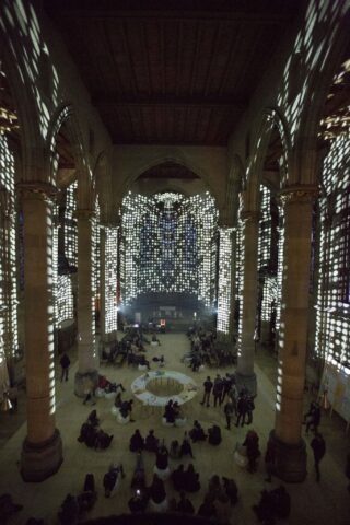 In einer großen Kirche mit hohen Säulen sitzen Menschen auf dem Boden, während gemusterte Lichter auf die Wände und die Decke projiziert werden und so im gesamten Raum einen gepunkteten Effekt erzeugen.
