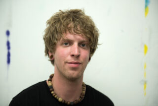 Ein junger Mann mit hellbraunem, lockigem Haar, einem schwarzen Hemd, einer Halskette aus Holzperlen und Ohrstöpseln posiert vor einem schlichten weißen Hintergrund mit schwachen Farbmarkierungen.