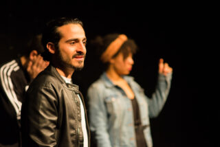 Drei Personen stehen in einem schwach beleuchteten Raum. Der Mann vorne trägt eine schwarze Lederjacke, während zwei weitere im Hintergrund leicht unscharf sind.