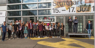 Eine Gruppe von Menschen steht vor einem Gebäude mit der Aufschrift „NEUBAU 2“, während mehrere Personen auf einem Podium zu ihnen sprechen; im Hintergrund ist ein großes Veranstaltungsbanner zu sehen.