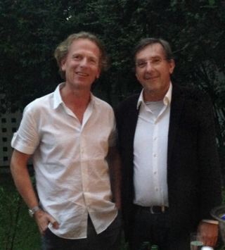 Zwei Männer stehen nebeneinander im Freien. Beide tragen Hemden mit Kragen. Einer trägt ein weißes Kurzarmhemd, der andere ein weißes Hemd und eine dunkle Jacke. Im Hintergrund sind Bäume und ein Zaun zu sehen.