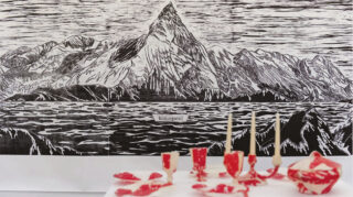 Hinter einem weißen Tisch mit rotem und weißem Geschirr und drei brennenden Stabkerzen ist ein schwarz-weißes Kunstwerk mit einer Berglandschaft ausgestellt.