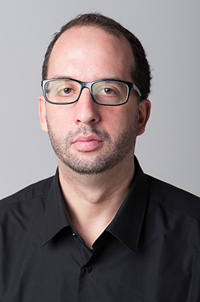 Ein Mann mit kurzen dunklen Haaren, Brille und gestutztem Bart trägt ein schwarzes Hemd mit Kragen und blickt vor einem schlichten grauen Hintergrund in die Kamera.