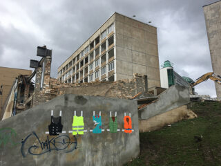 Teilweise abgerissenes Gebäude mit Baumaschinen im Hintergrund; Graffiti und bunte Westenillustrationen auf einer Betonwand im Vordergrund.