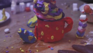 Ein roter Becher aus Modelliermasse ist mit buntem Ton gefüllt, umgeben von verschiedenen bunten Tonformen und verstreuten kleinen Objekten auf einer Holzoberfläche.