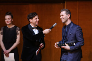 Ein Mann im Anzug wird auf der Bühne mit einem Mikrofon interviewt, während er eine Auszeichnung in der Hand hält; zwei weitere Personen stehen in der Nähe, eine davon hält ein Papier in der Hand.