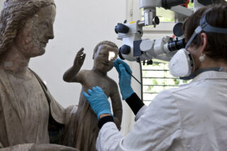 Ein Restaurator mit Handschuhen und Maske restauriert eine Statue einer Frau und eines Kindes mithilfe von Präzisionswerkzeugen und einem Mikroskop.