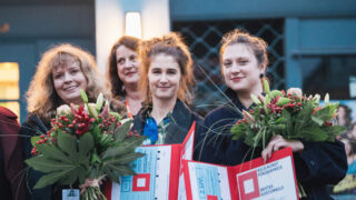 Vier Frauen stehen mit Blumensträußen und Urkunden in den Händen zusammen und posieren für ein Foto bei einer Veranstaltung, die wie eine Preisverleihung aussieht.