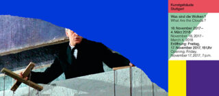 Hinter einer Betonbarriere steht ein Mann im Anzug und hält zwei Holzstäbe in der Hand. Auf einem bunten Block rechts davon werden Veranstaltungsdetails auf Deutsch und Englisch angezeigt.