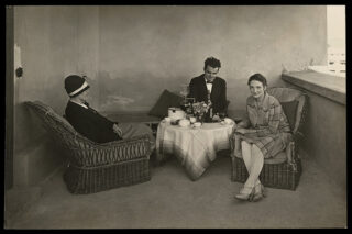 Drei Personen sitzen mit Tee und Blumen um einen kleinen Tisch auf einer Terrasse. Zwei Frauen tragen Kleider, ein Mann trägt einen Anzug. Das Ambiente wirkt formell und nostalgisch.
