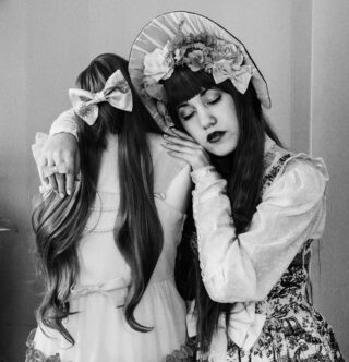 Eine Frau in Vintage-Kleidung mit einer geblümten Haube umarmt eine kopflose Schaufensterpuppe, die ein Kleid und eine lange Perücke mit einer großen Schleife trägt.