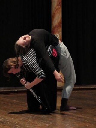 Zwei Personen treten auf der Bühne auf; eine spielt Klarinette, während sie nach vorne gebeugt ist, die andere lehnt sich mit geschlossenen Augen über sie. Beide stehen barfuß auf einem Holzboden vor dunklem Hintergrund.