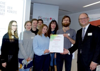 Eine Gruppe von sieben Personen steht drinnen, einige lächeln, während ein Mann im Anzug einem Mann mit Bart und Brille eine Auszeichnungsurkunde überreicht.