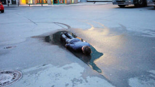 Eine Person liegt mit dem Gesicht nach unten in einer großen Pfütze auf einer leeren Stadtstraße, die sich teilweise im Wasser spiegelt, während umliegende Gebäude und Fahrzeuge sichtbar sind.